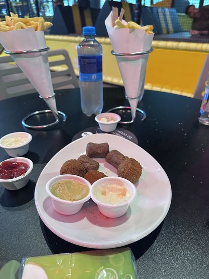 A table setting featuring two cone-shaped servings of fries, a bottle of water, and a plate with several fried snacks accompanied by three small dipping sauce containers (two white sauces and one red sauce).